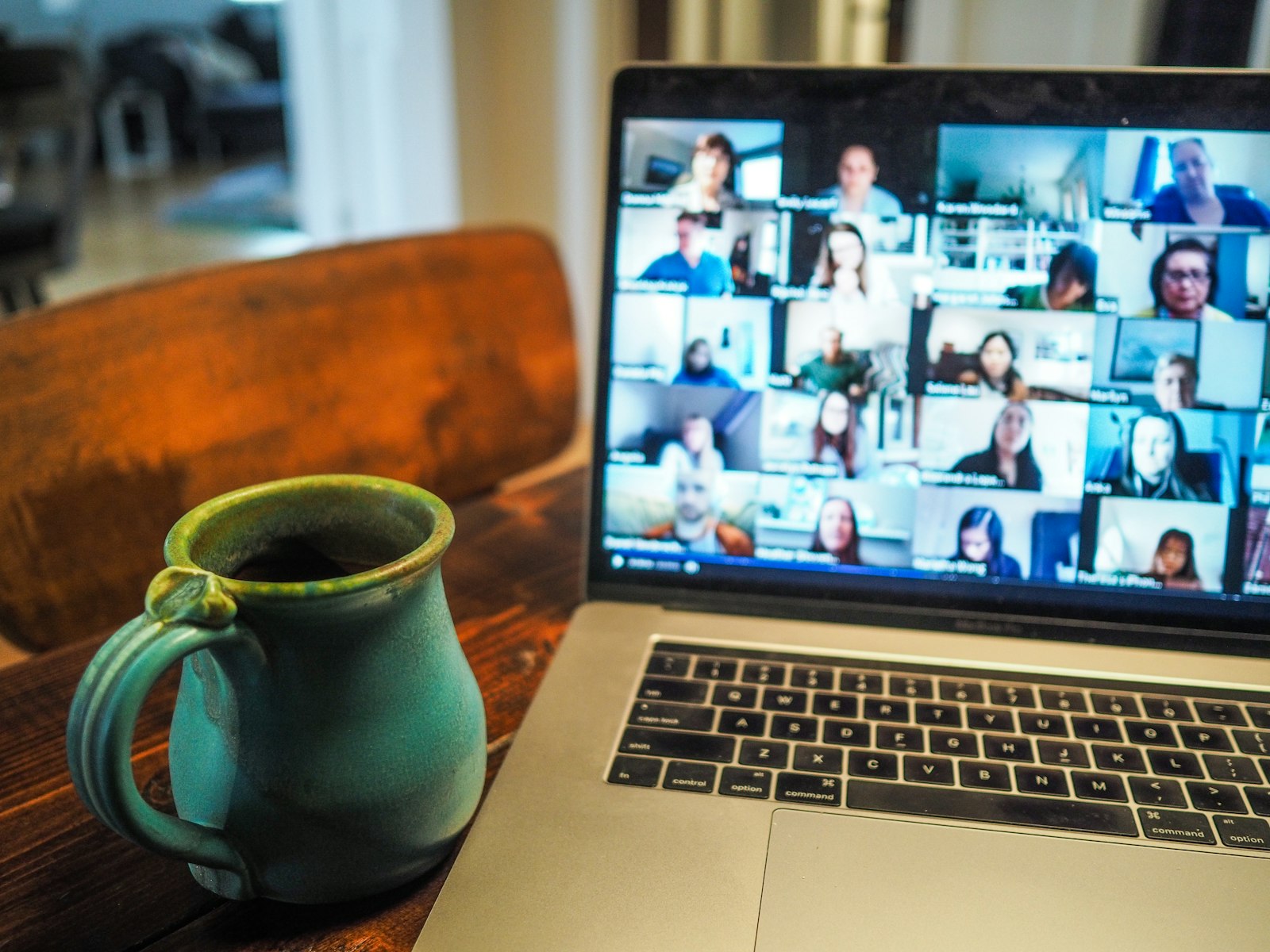 A laptop on a wooden table showing a video call with a grid of colleagues, beside a green mug